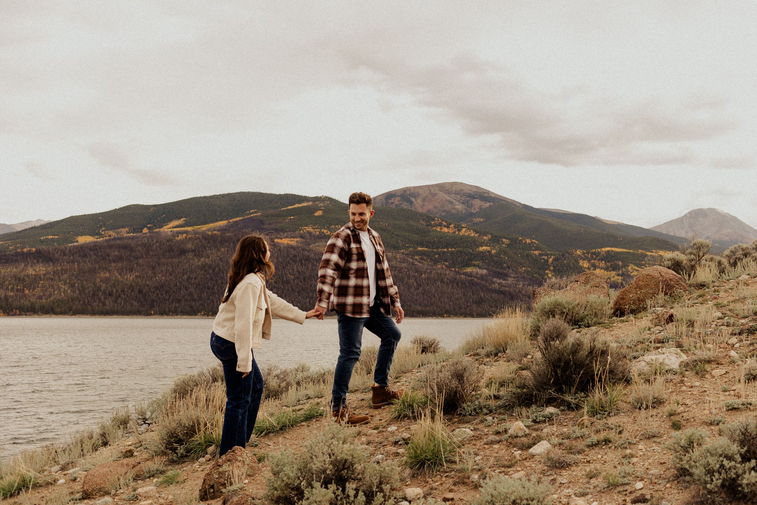 Couple holding hands on a rocky lakeside trail with mountain views and cloudy skies in a scenic outdoor setting.