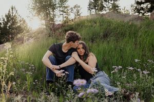 Couple sits together in a wildflower meadow at golden hour, sharing a tender romantic moment outdoors.