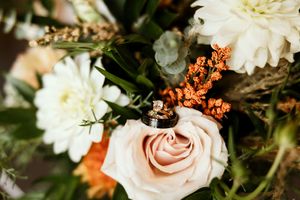 Wedding rings rest on a blush pink rose surrounded by white dahlias and orange berries in a bridal bouquet.