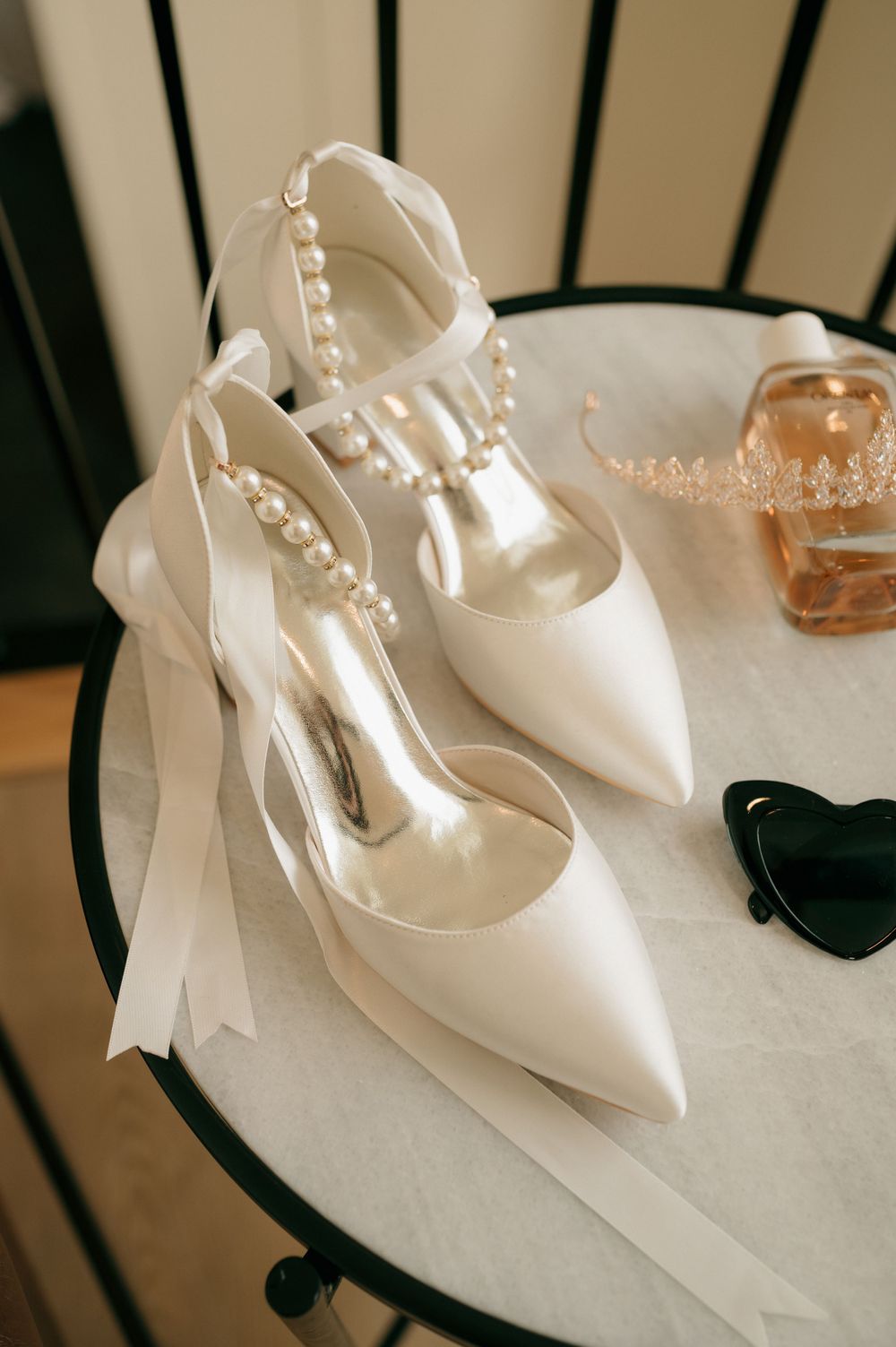 Elegant white bridal shoes with pearl details and ribbons displayed on a round table.