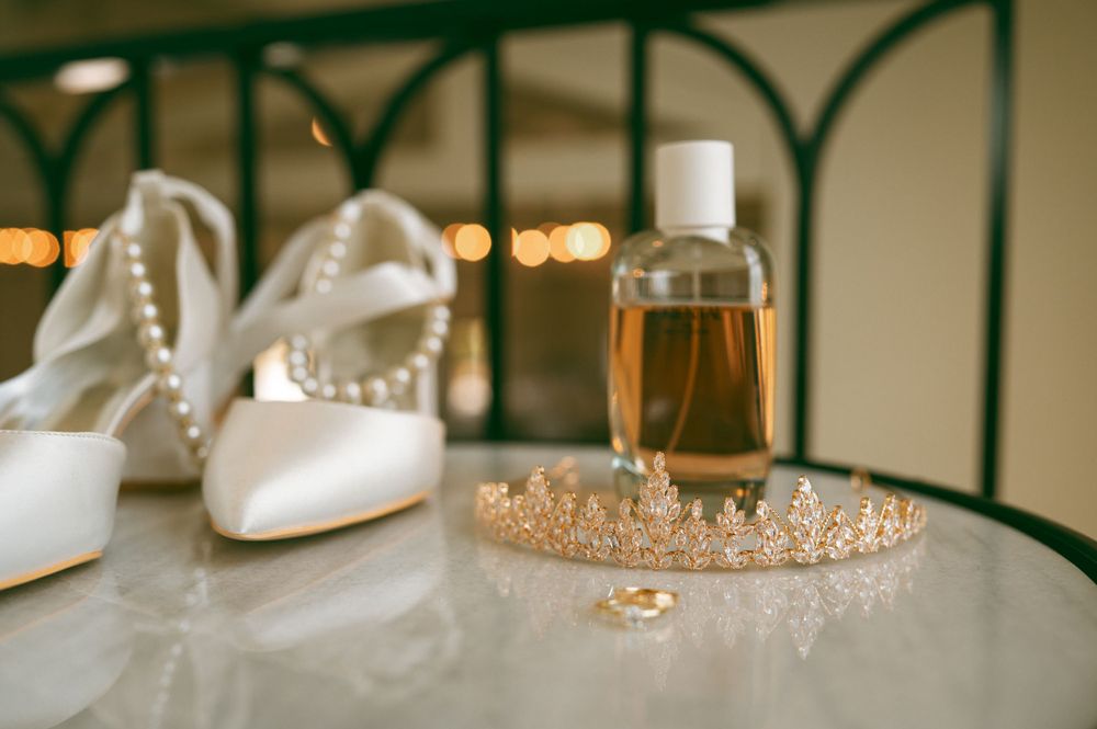 Wedding accessories including white heels, crystal tiara and champagne glass arranged on reflective surface.