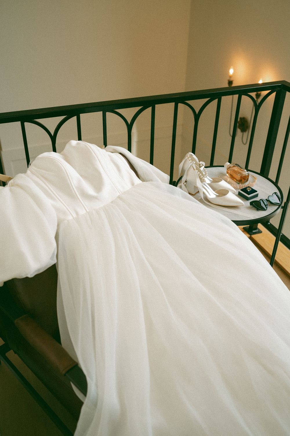 A white wedding dress laid out on a bed with green metal headboard and white shoes on a side table.