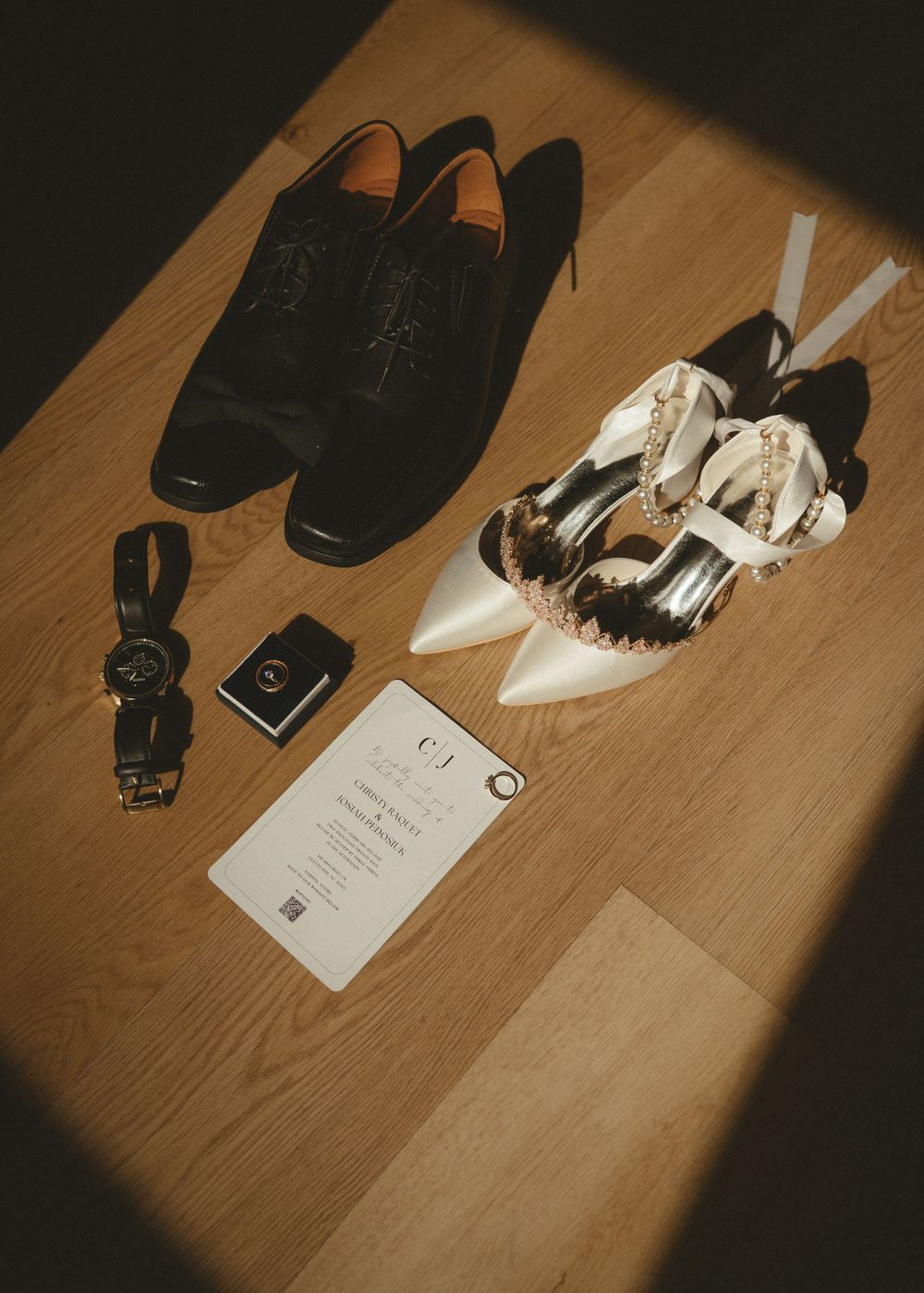 Wedding day details with formal shoes, accessories and invitation laid out on wooden floor.