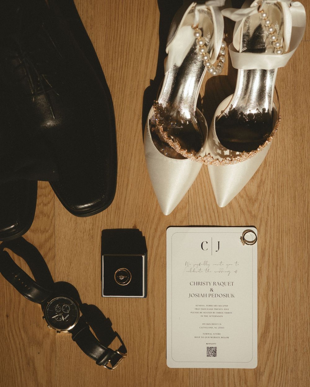 Wedding details flatlay with white heels, black shoes, rings and invitation card on wooden floor.