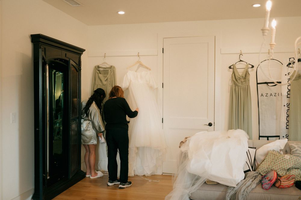 Two people looking at a white wedding dress displayed on a hanger in a bridal fitting room with a mirror and furniture.