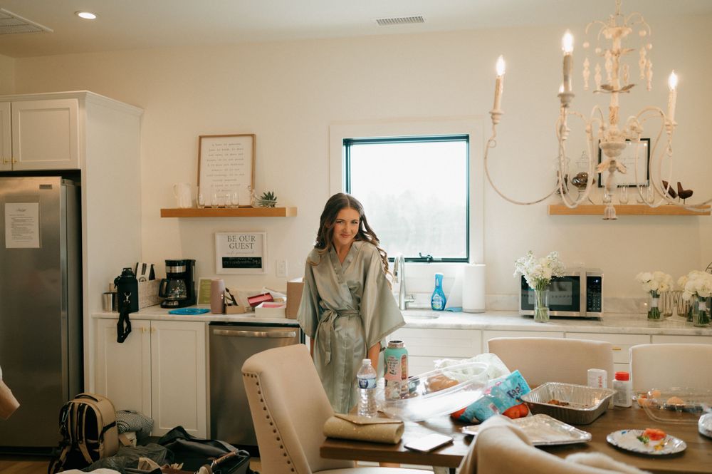 A woman in a robe stands in a stylish white kitchen with candles and catering items on the counter.