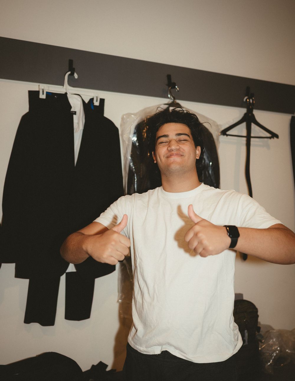 A person giving thumbs up while posing next to a formal black suit hanging on the wall.