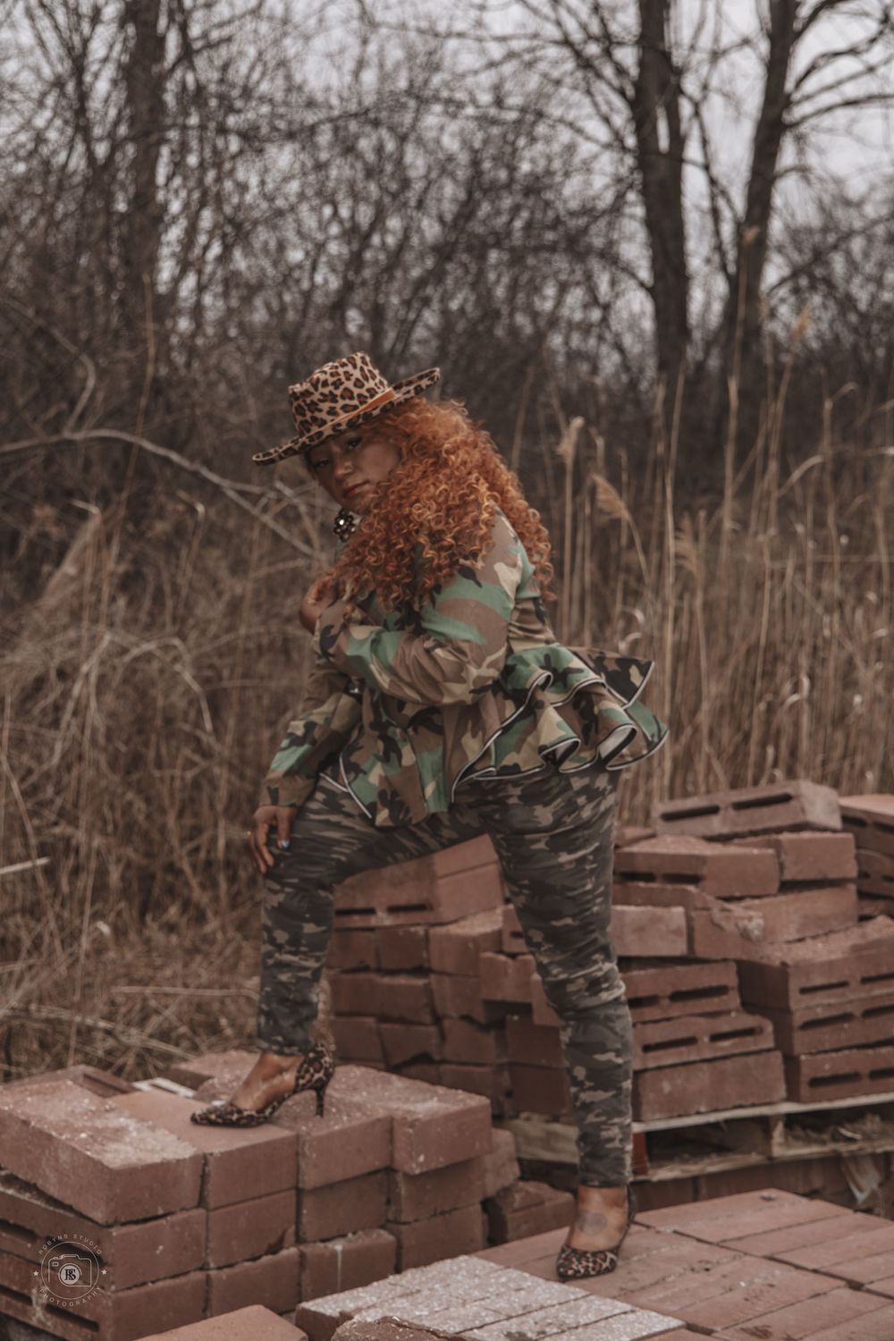 Woman in camo outfit and leopard hat poses on stacked bricks in an outdoor setting with bare trees.