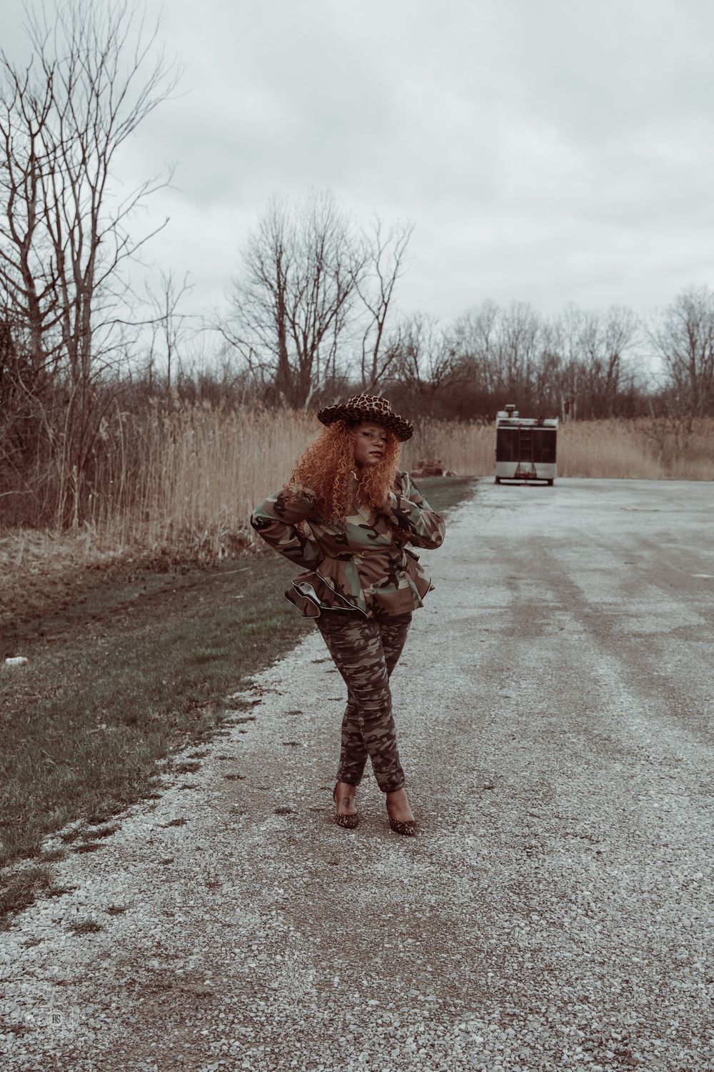 Woman in camouflage outfit and leopard print hat posing on a rural gravel road surrounded by bare trees.