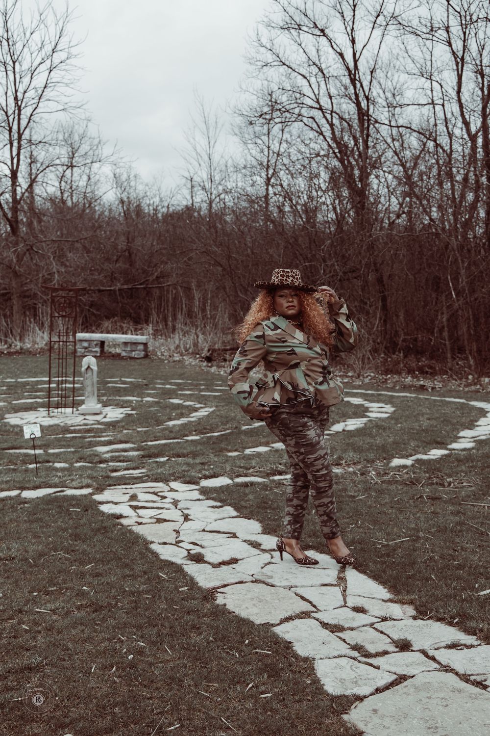 Woman in camouflage outfit and leopard hat poses on stone path in bare winter garden landscape.