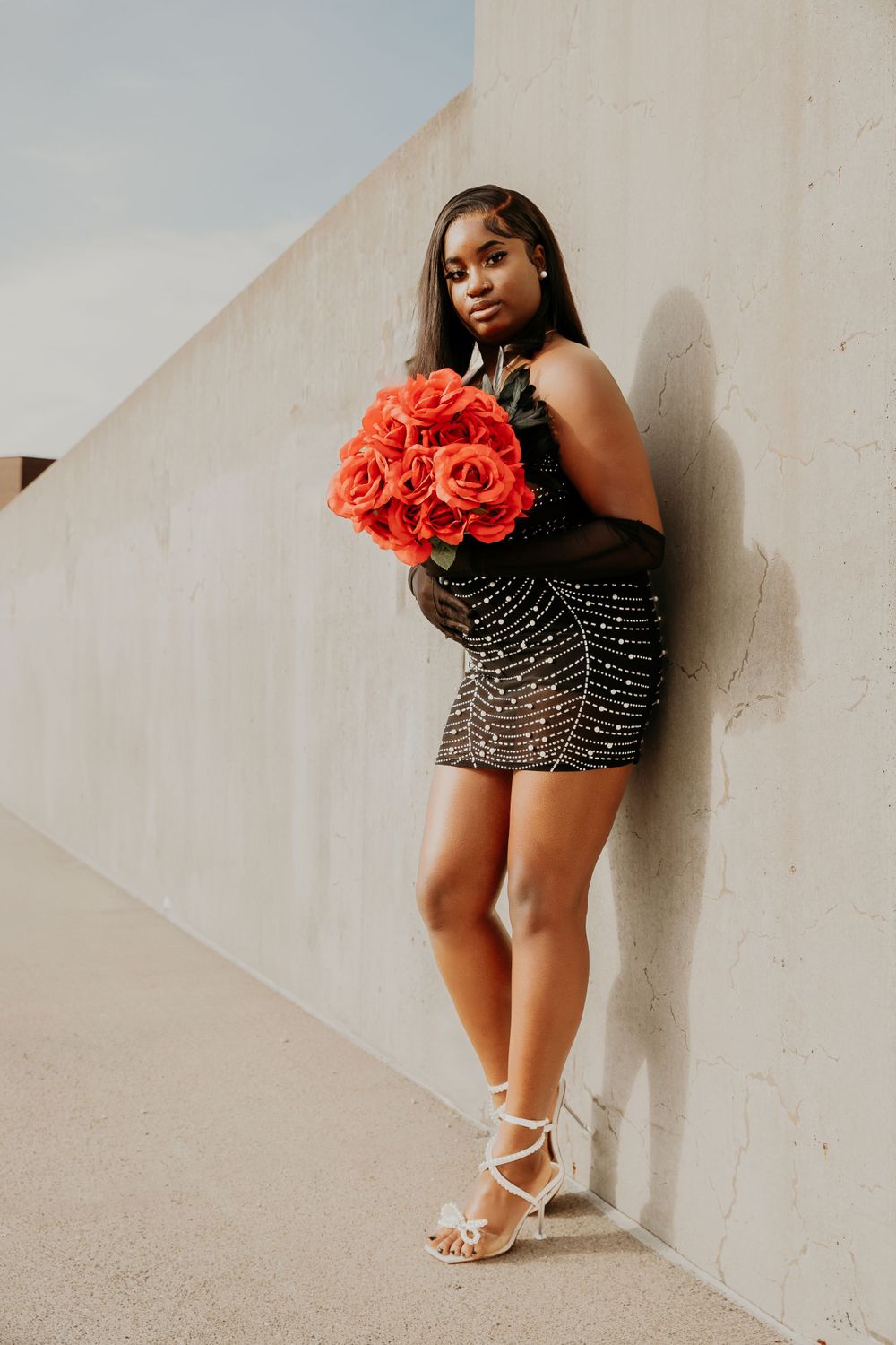Figure in a sparkly dress poses with red flowers against a light wall.