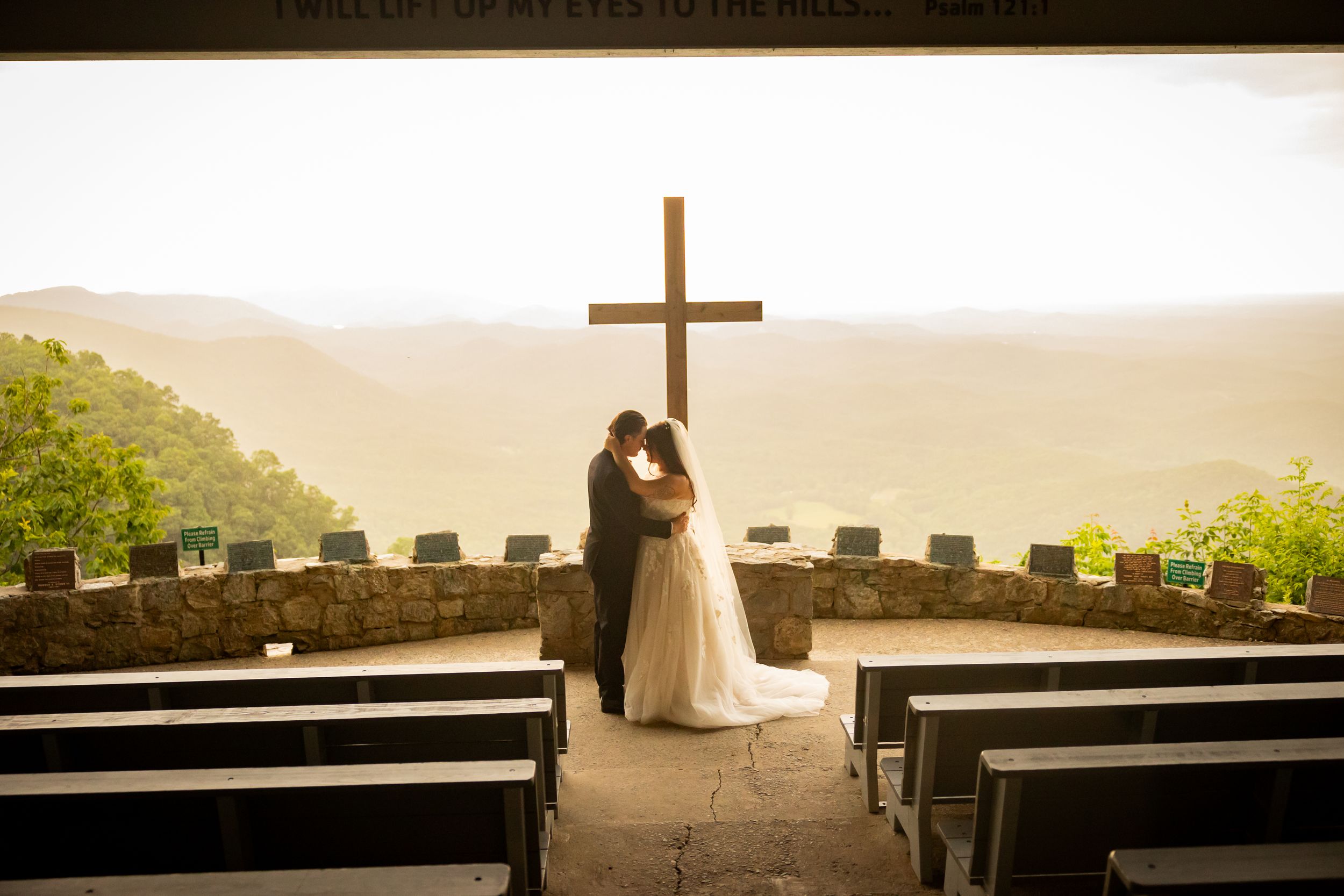 A couple stands as silhouettes at an outdoor mountain chapel with a cross and empty wooden pews at sunset.