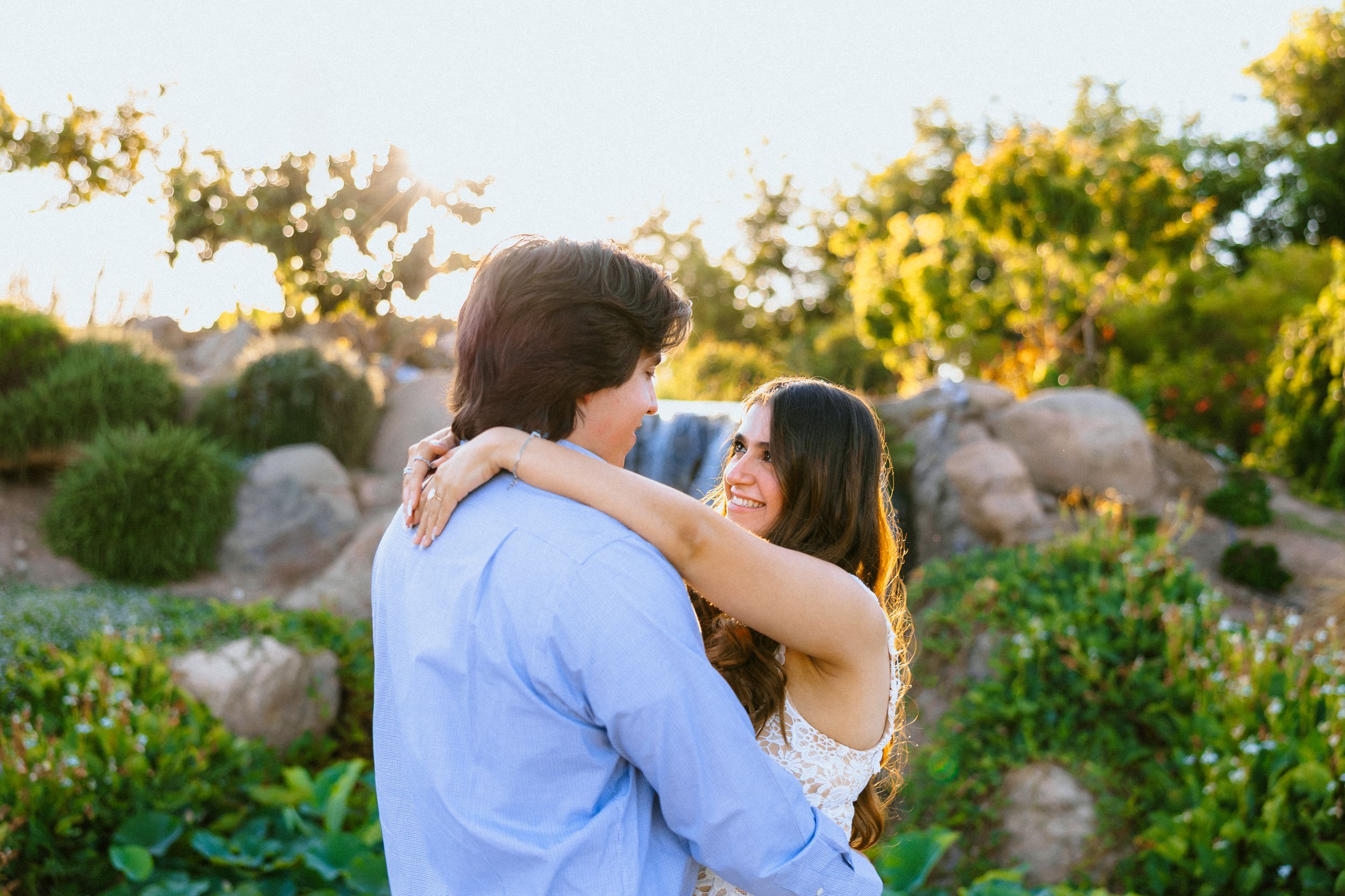 Couple embracing in a sunlit garden, woman smiling over man's shoulder amid lush greenery and rocks.