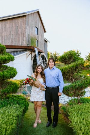 Couple posing in lush garden path before rustic barn with water wheel, woman holding bouquet in white dress.