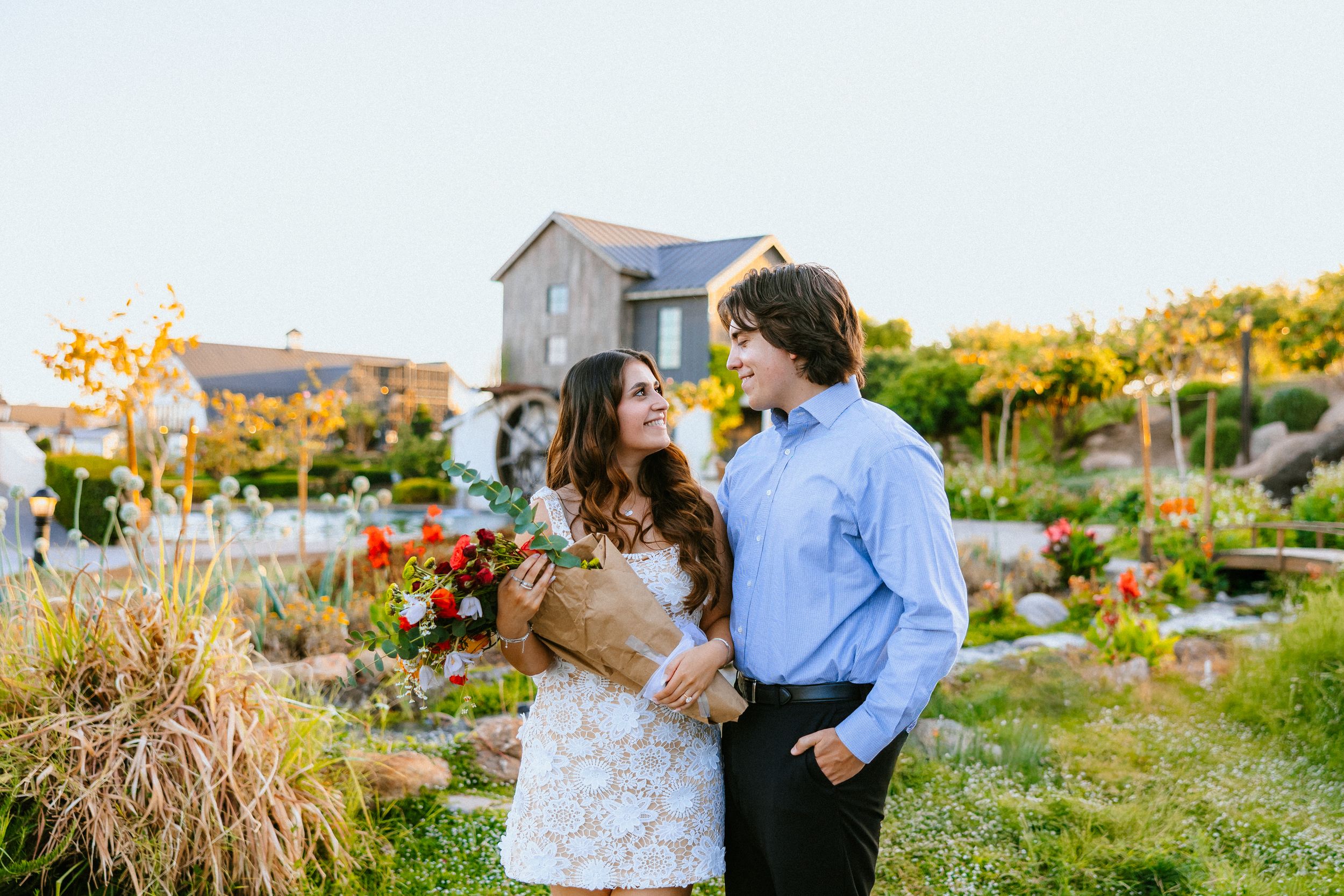 Couple embracing in a garden, woman in white lace dress holding a bouquet, man in blue shirt, with a rustic barn backdrop.