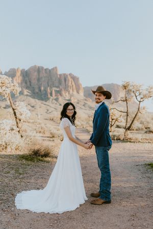 Bride and groom hold hands in desert landscape with Superstition Mountains backdrop, she in white gown, he in cowboy hat.