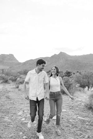 A couple walks together smiling on a desert trail with mountains in the background.