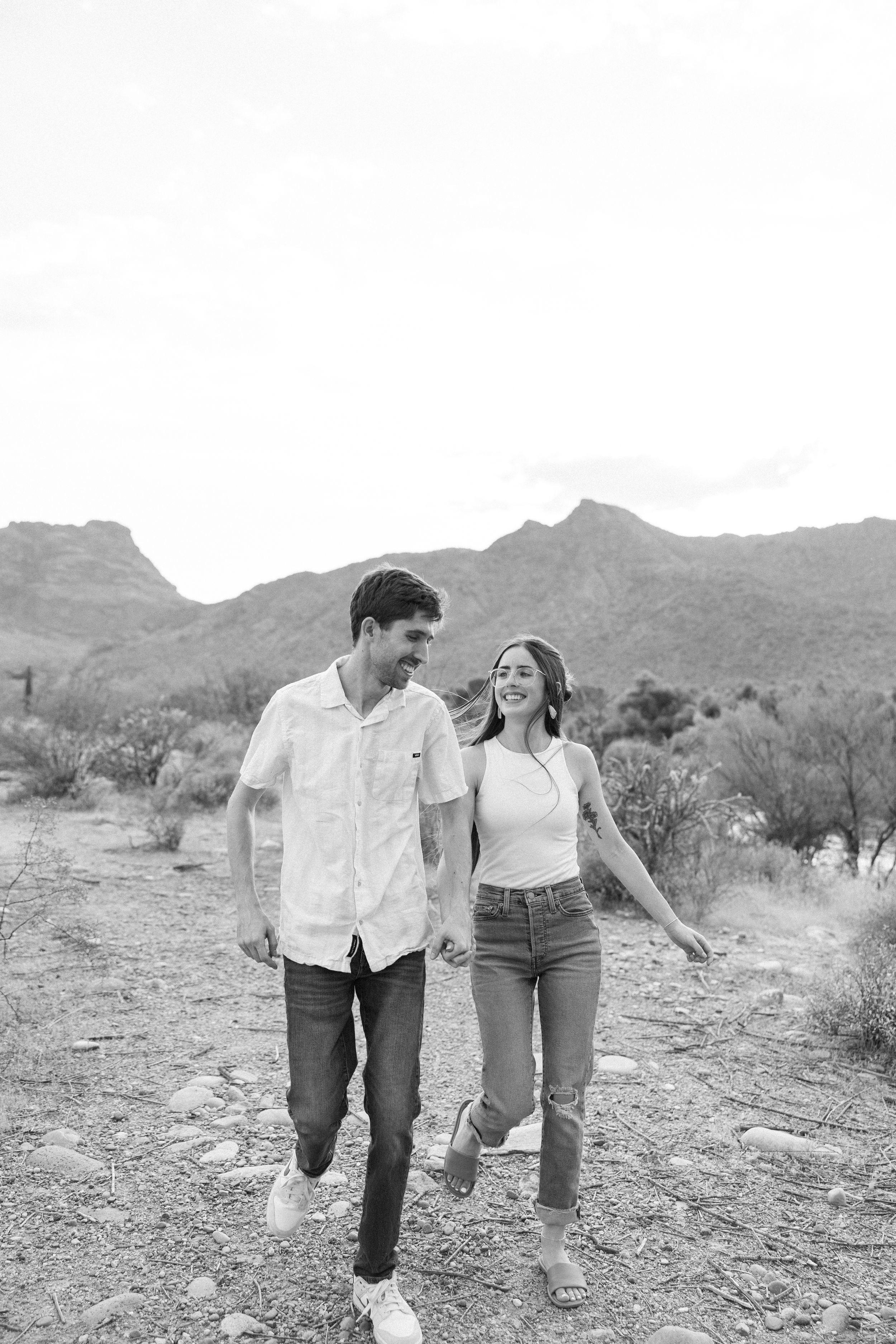 A couple walks together smiling on a desert trail with mountains in the background.