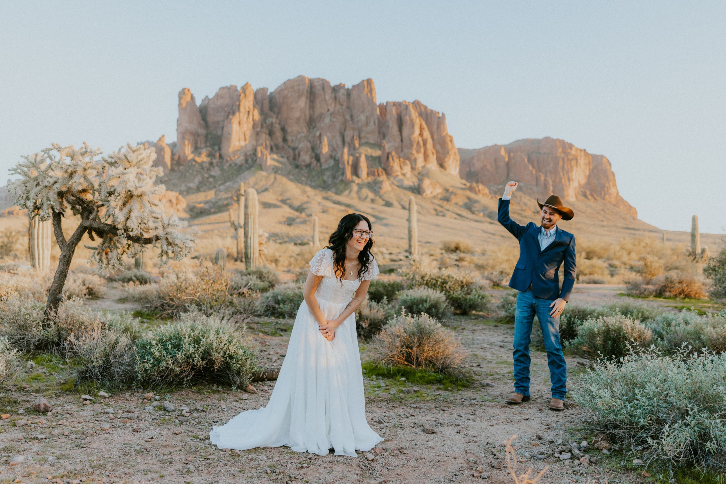 Couple in wedding attire poses in Arizona desert with Superstition Mountains backdrop at golden hour.