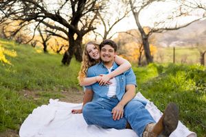 Couple sitting on white blanket in green field with bare trees at golden hour sunset.