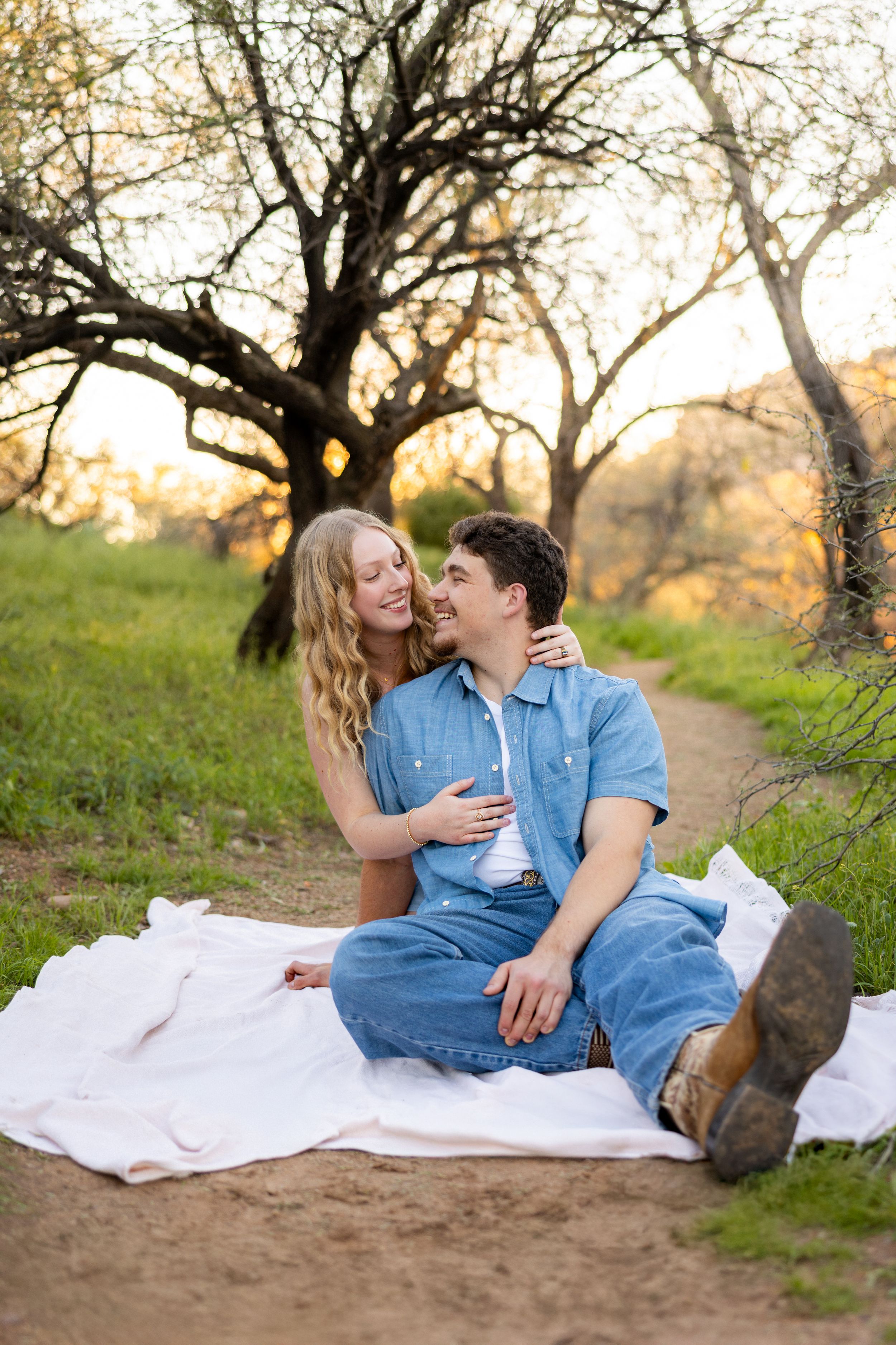 Couple sitting on white blanket outdoors at golden hour, woman embracing man from behind, surrounded by trees and green grass.