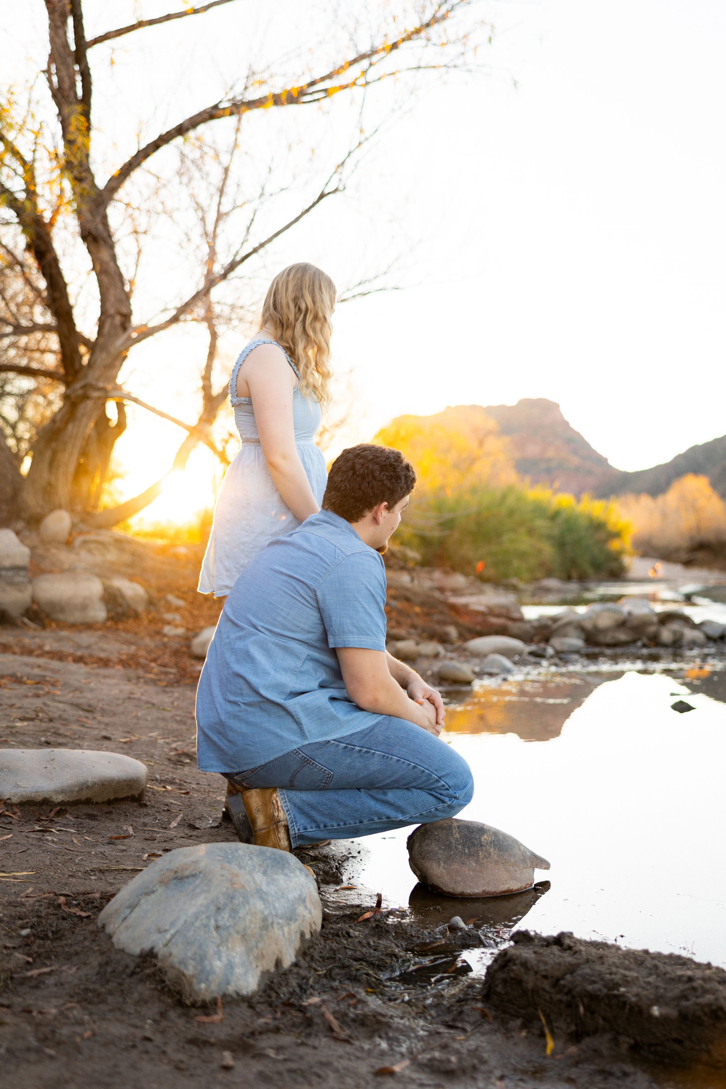 Couple by a rocky riverside at golden sunset, man kneeling beside woman in white dress near tranquil water.