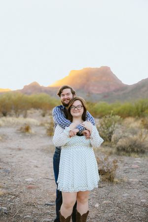 Couple embracing at golden hour in desert landscape with mountain backdrop, woman in white dress and cowboy boots.