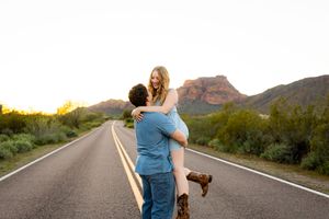 Man lifting woman on empty desert road at sunset with mountain backdrop, romantic couple photography.