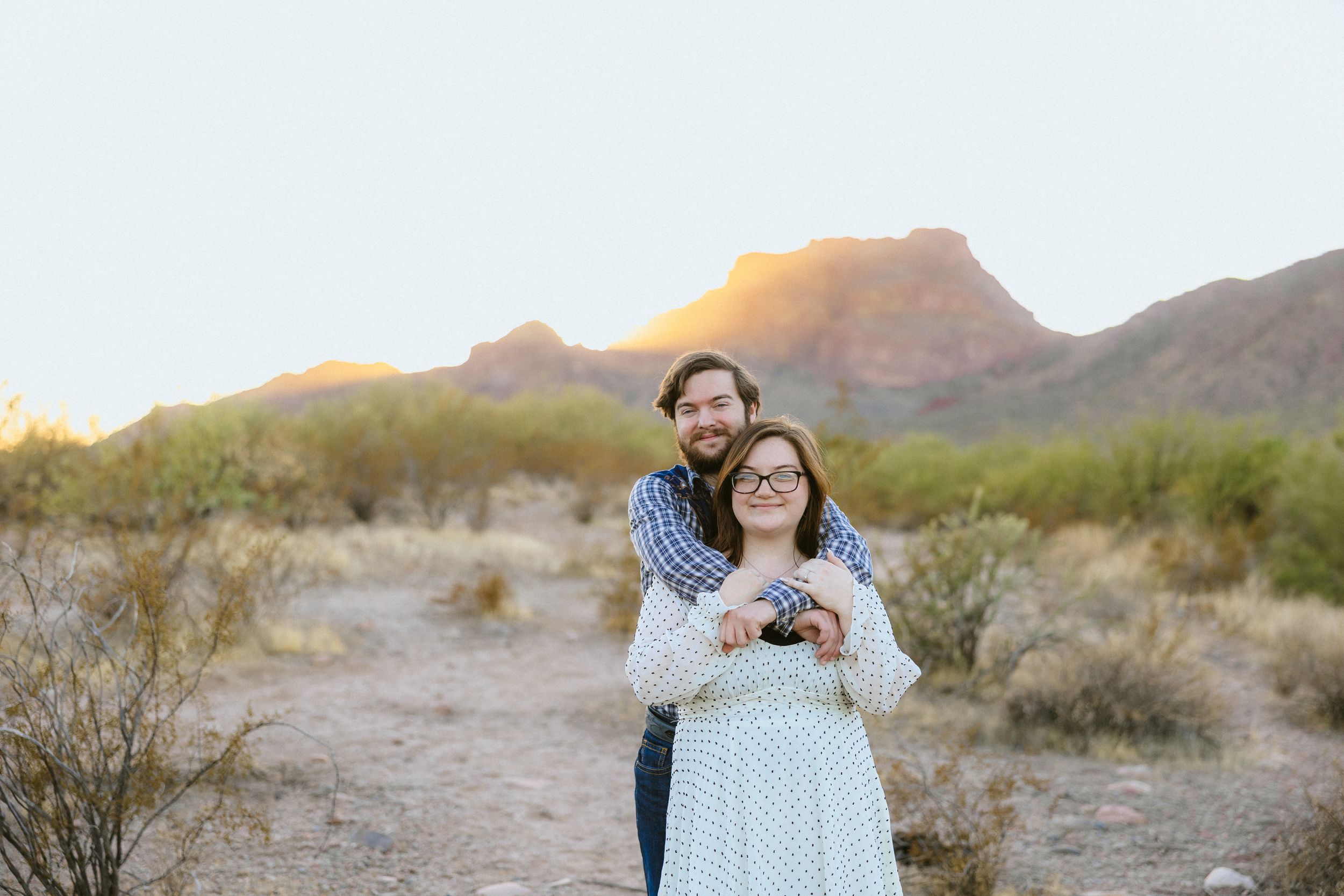 Couple embracing in desert landscape with mountain backdrop at golden hour sunset in Arizona.