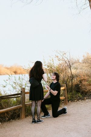 Man proposing on one knee to woman on a scenic outdoor path by a lake surrounded by autumn trees.