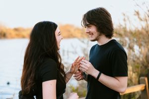 Young couple in black shirts holding hands and smiling at each other near a lake at golden hour.