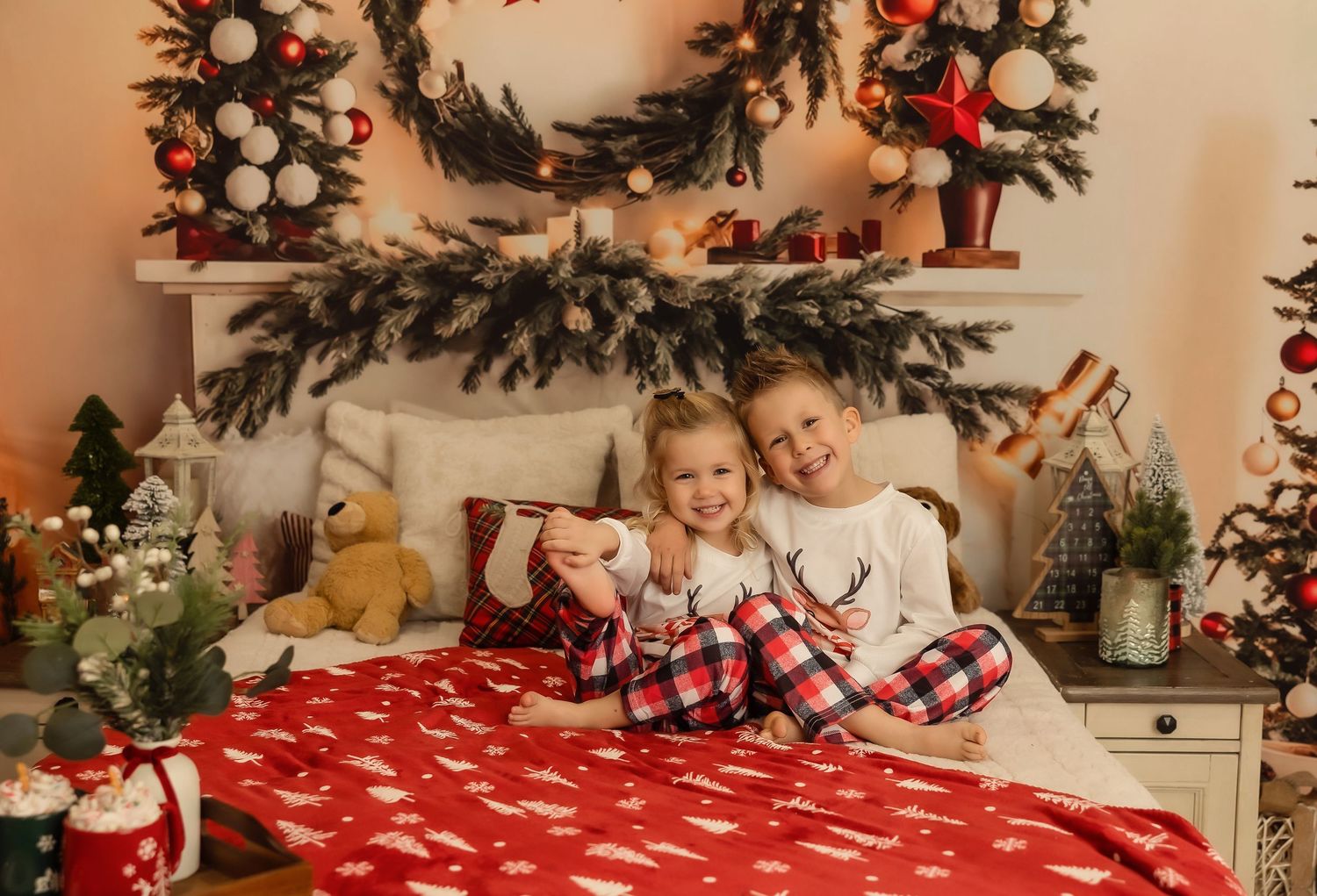 Two children in Christmas pajamas sitting on a red holiday bed surrounded by festive decorations and a wreath.