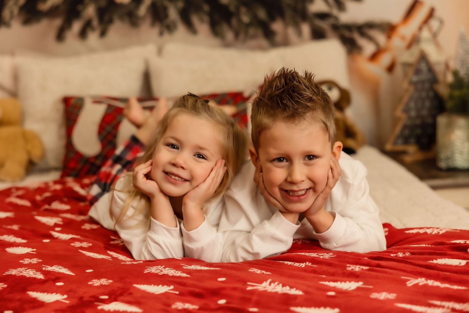 Two smiling children lying on a red Christmas blanket in festive holiday bedroom decor.