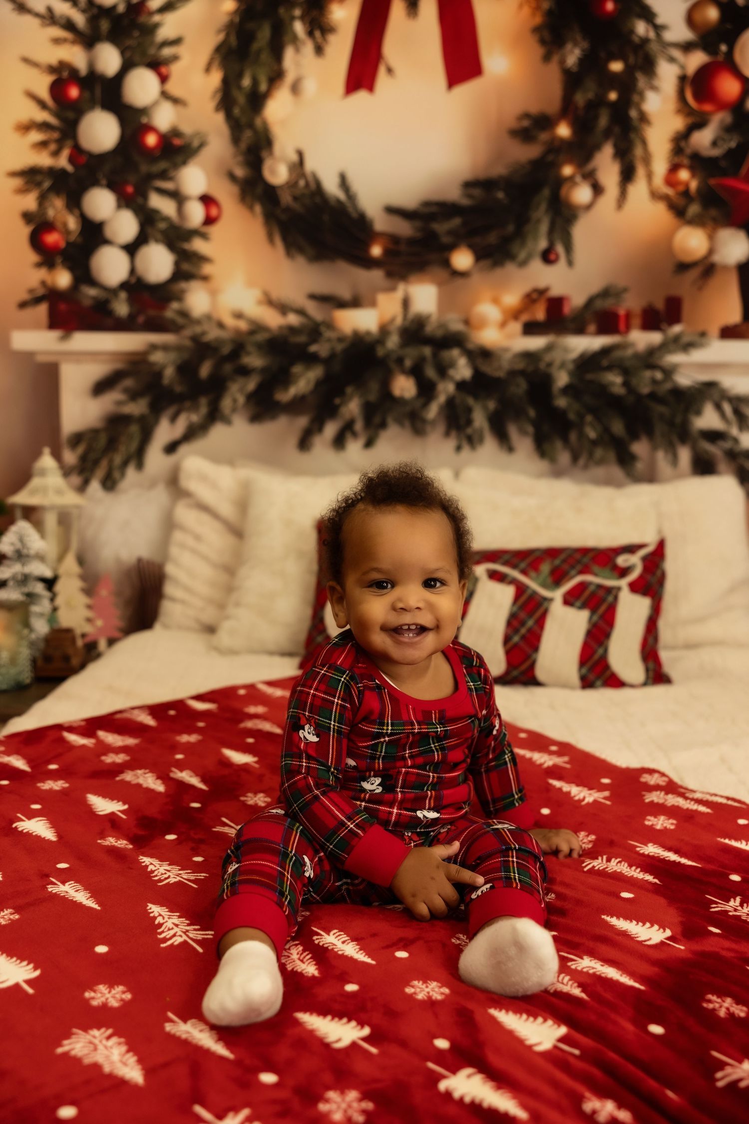Smiling baby in red plaid Christmas pajamas sitting on a festive red bed with holiday decorations in the background.