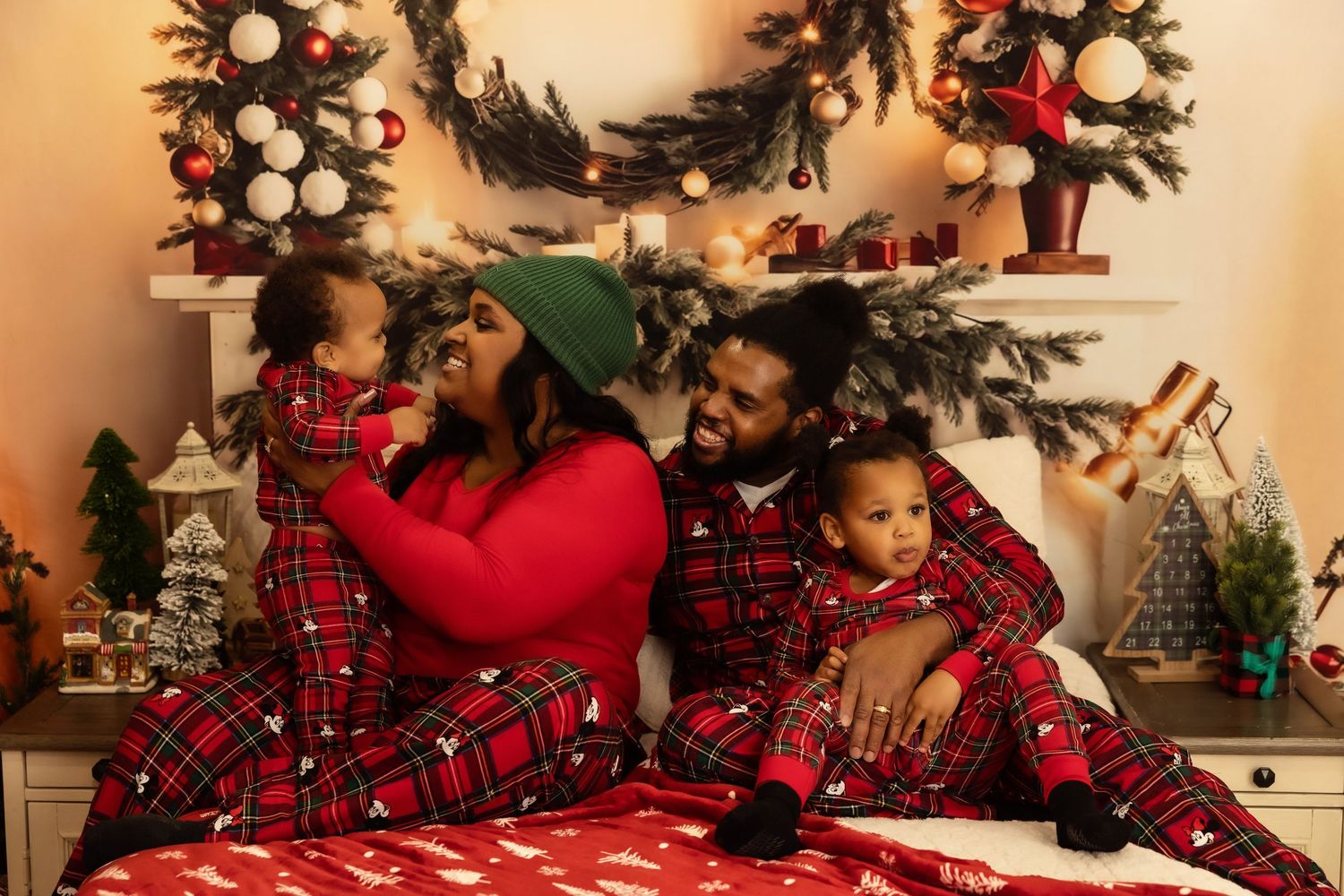 Family in matching red plaid Christmas pajamas sitting together on a bed with holiday decorations and a wreath backdrop.