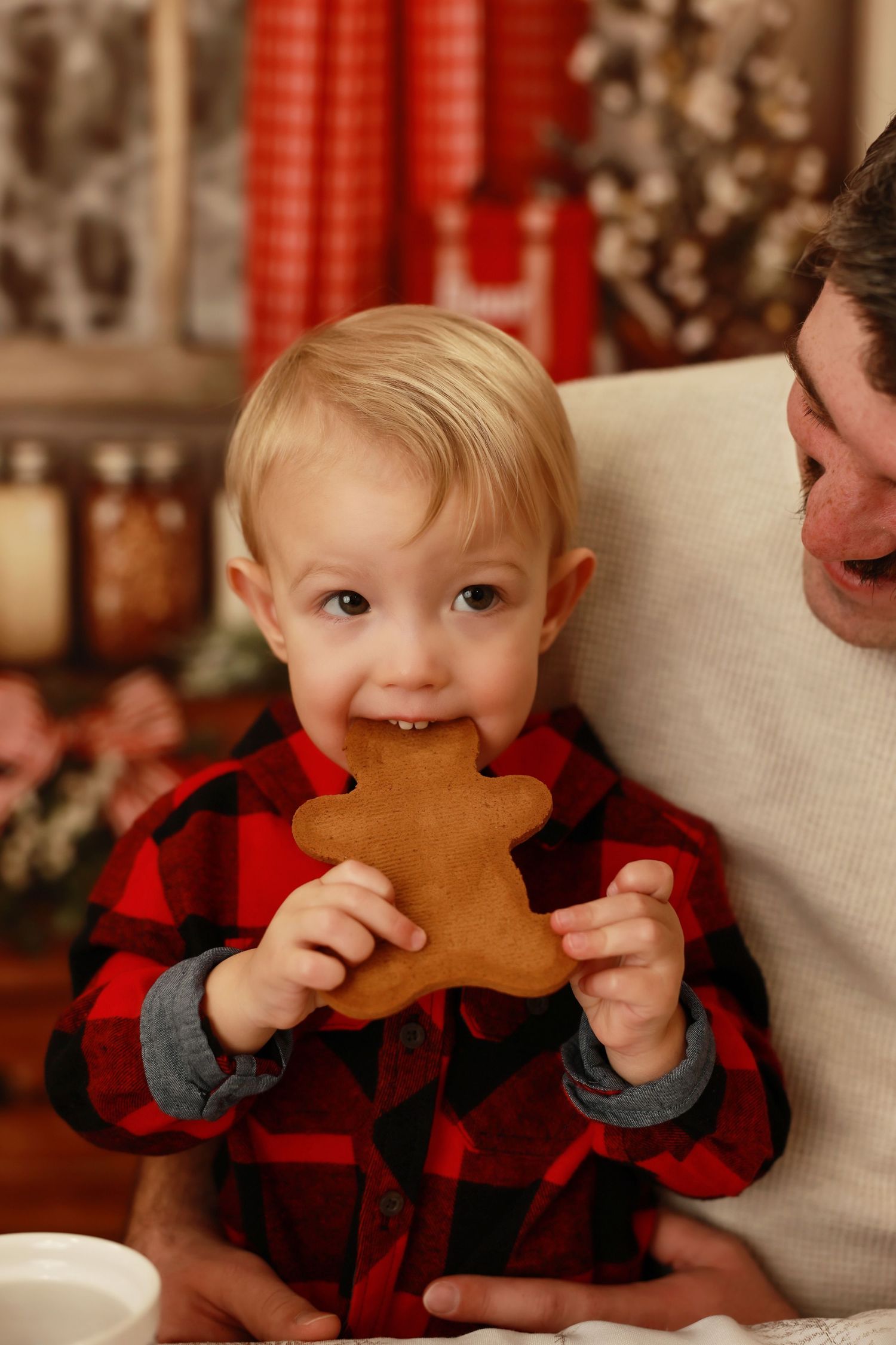 Toddler in red plaid shirt biting a gingerbread man cookie beside father in festive Christmas kitchen.