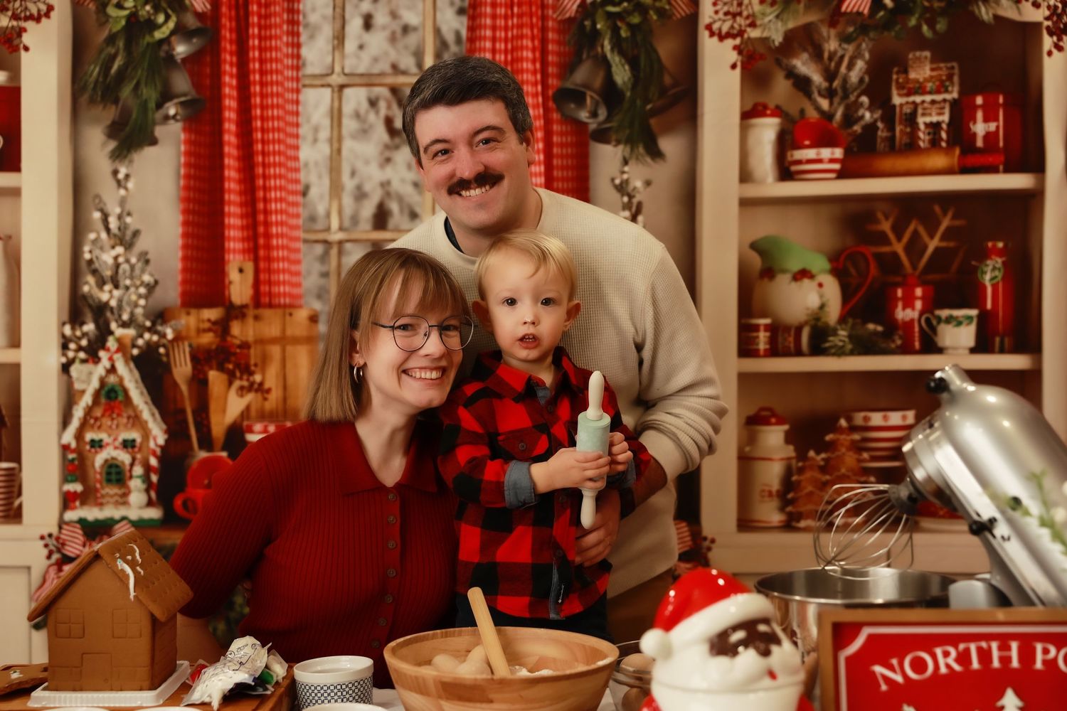 Family Christmas photo with parents and toddler in festive North Pole kitchen setting with holiday decorations.