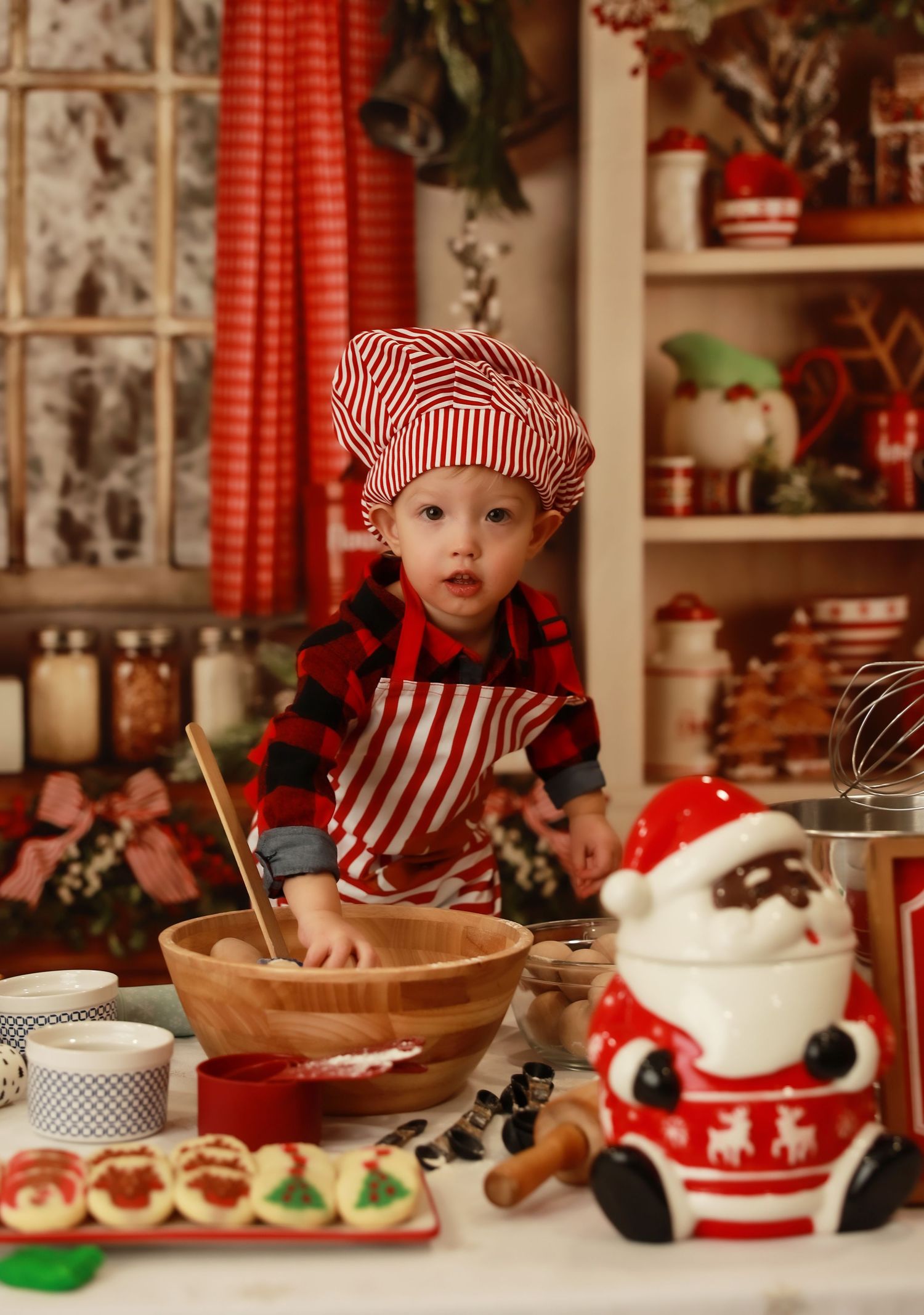 Toddler in red striped chef hat and apron mixing dough in wooden bowl with Santa cookie jar in Christmas kitchen.