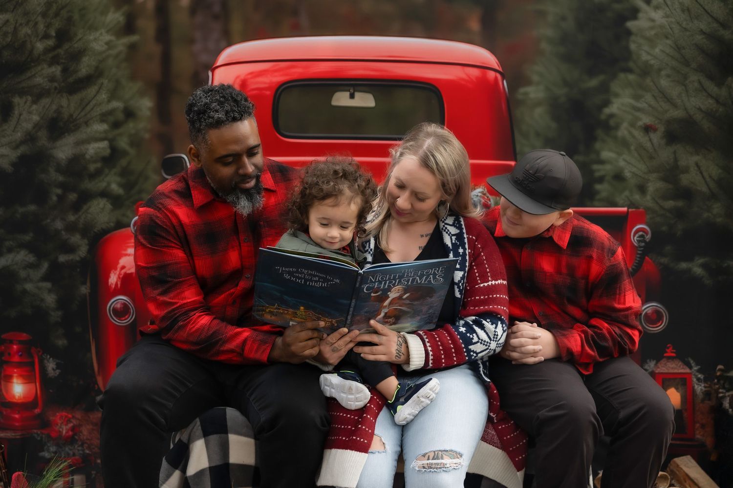 Family in matching red plaid shirts reading a Christmas book on a vintage red truck tailgate surrounded by pine trees.