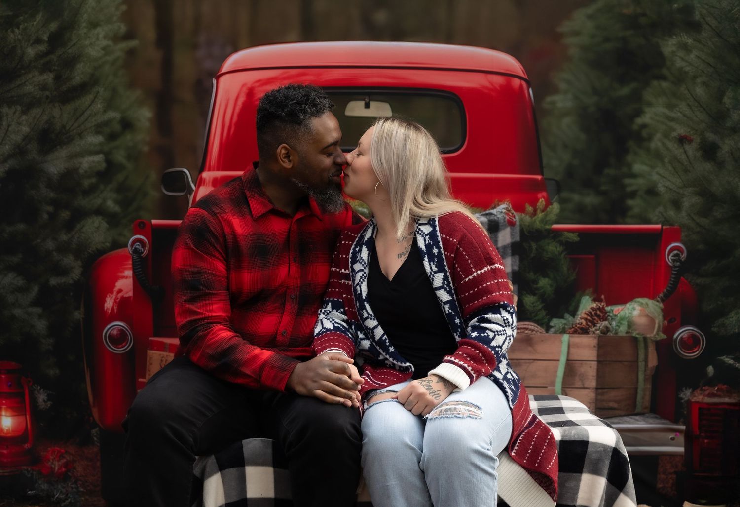 Couple kissing on red vintage truck bed surrounded by Christmas trees and holiday decorations.