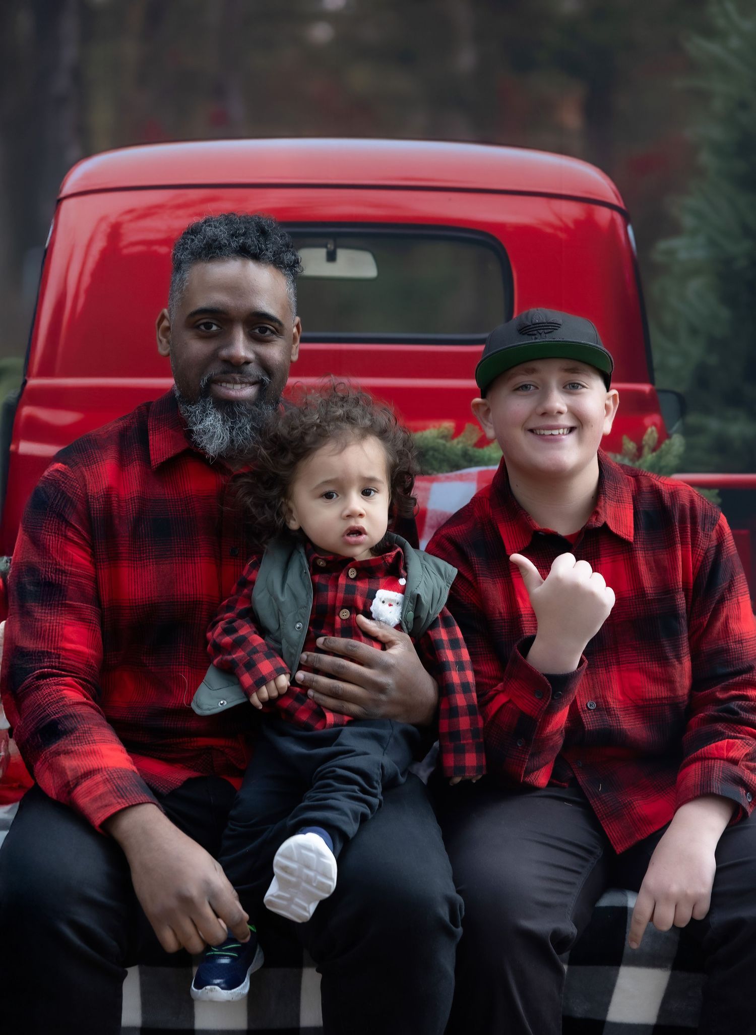 Three people in matching red plaid shirts sit on a red truck tailgate for a festive Christmas photo.