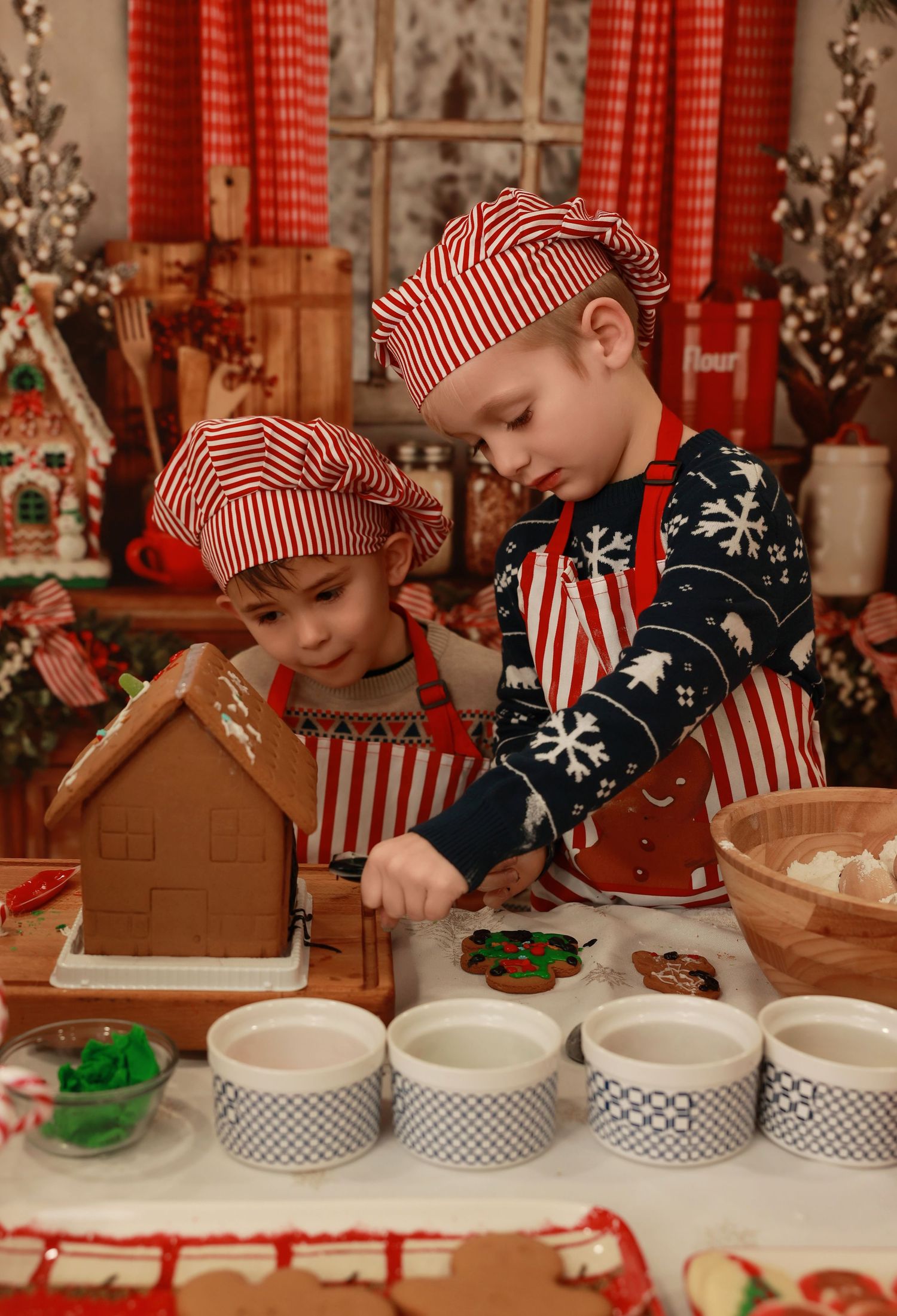 Two children in red striped chef hats and aprons decorating a gingerbread house at a festive Christmas table.