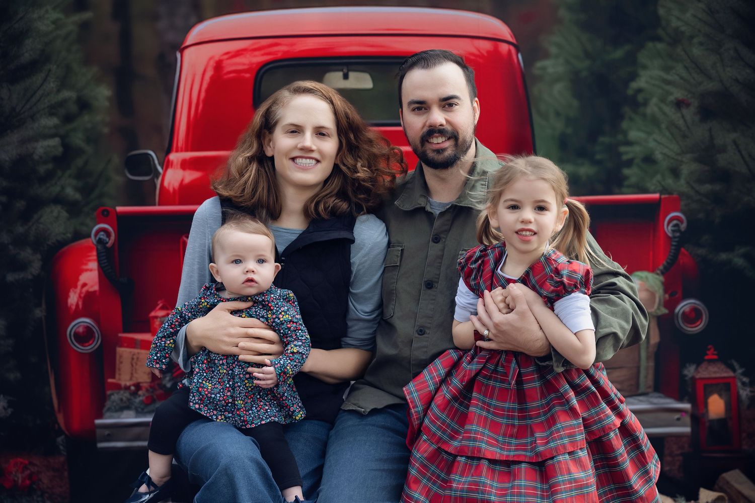 Family of four posing on red vintage truck tailgate surrounded by Christmas trees for holiday photo.