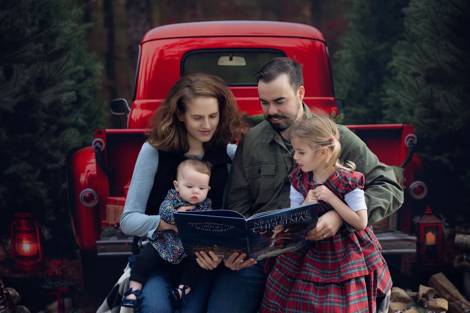 Family of four reading a Christmas book by a vintage red truck surrounded by pine trees.