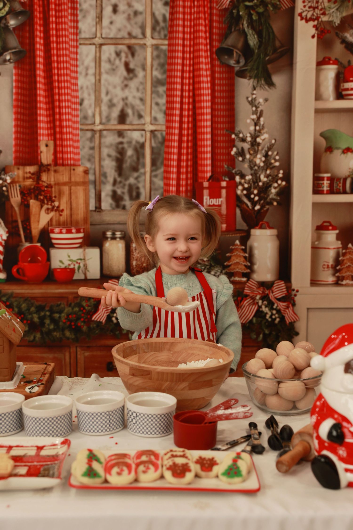 A cheerful toddler girl in a red apron bakes Christmas cookies in a festive holiday kitchen decorated with bells and garland.