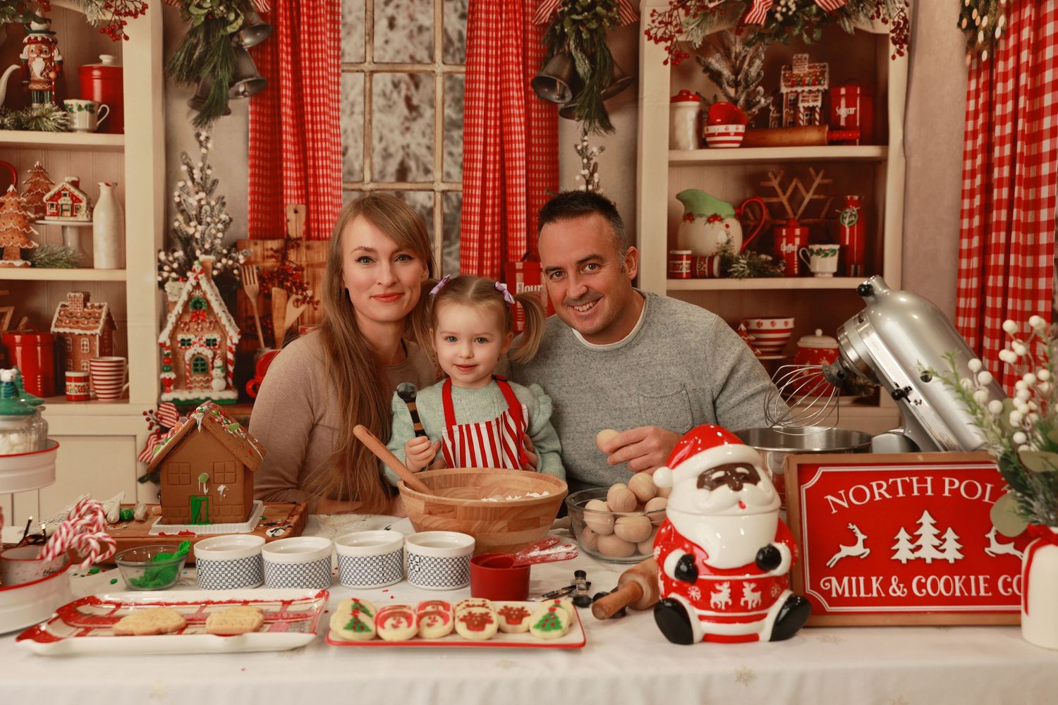 Family posing in festive Christmas kitchen with gingerbread house, cookies, and North Pole milk and cookie jar decor.