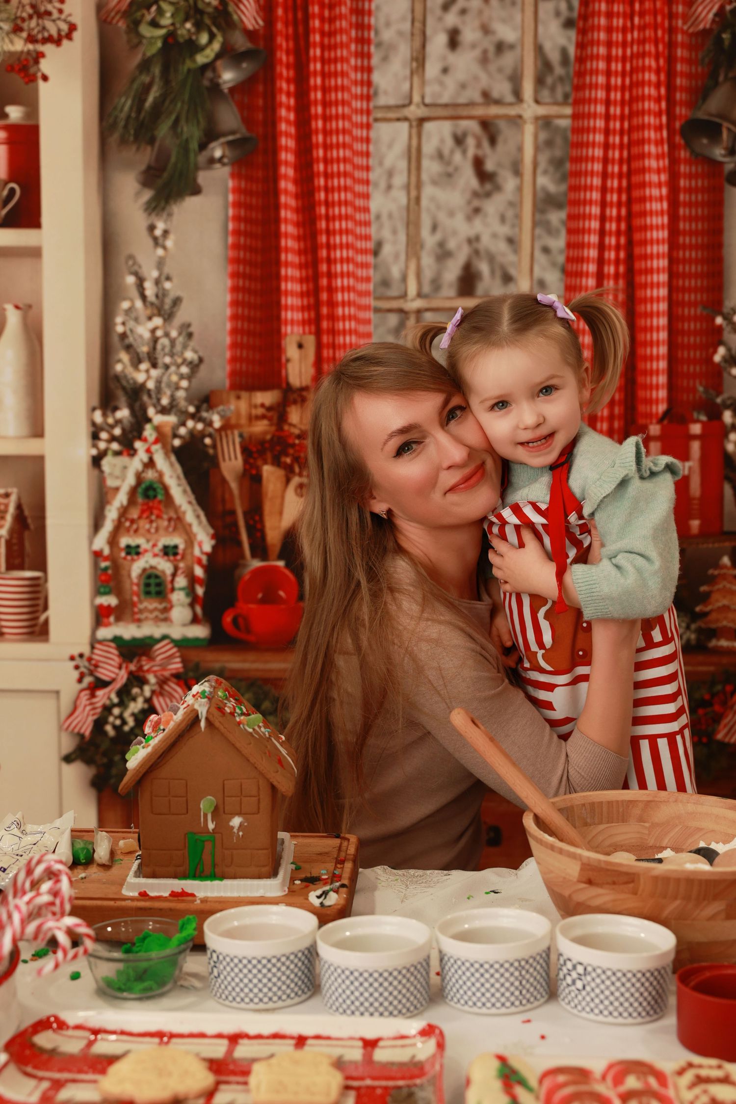 Mother and daughter baking Christmas cookies together in festive holiday kitchen with gingerbread house decorations.