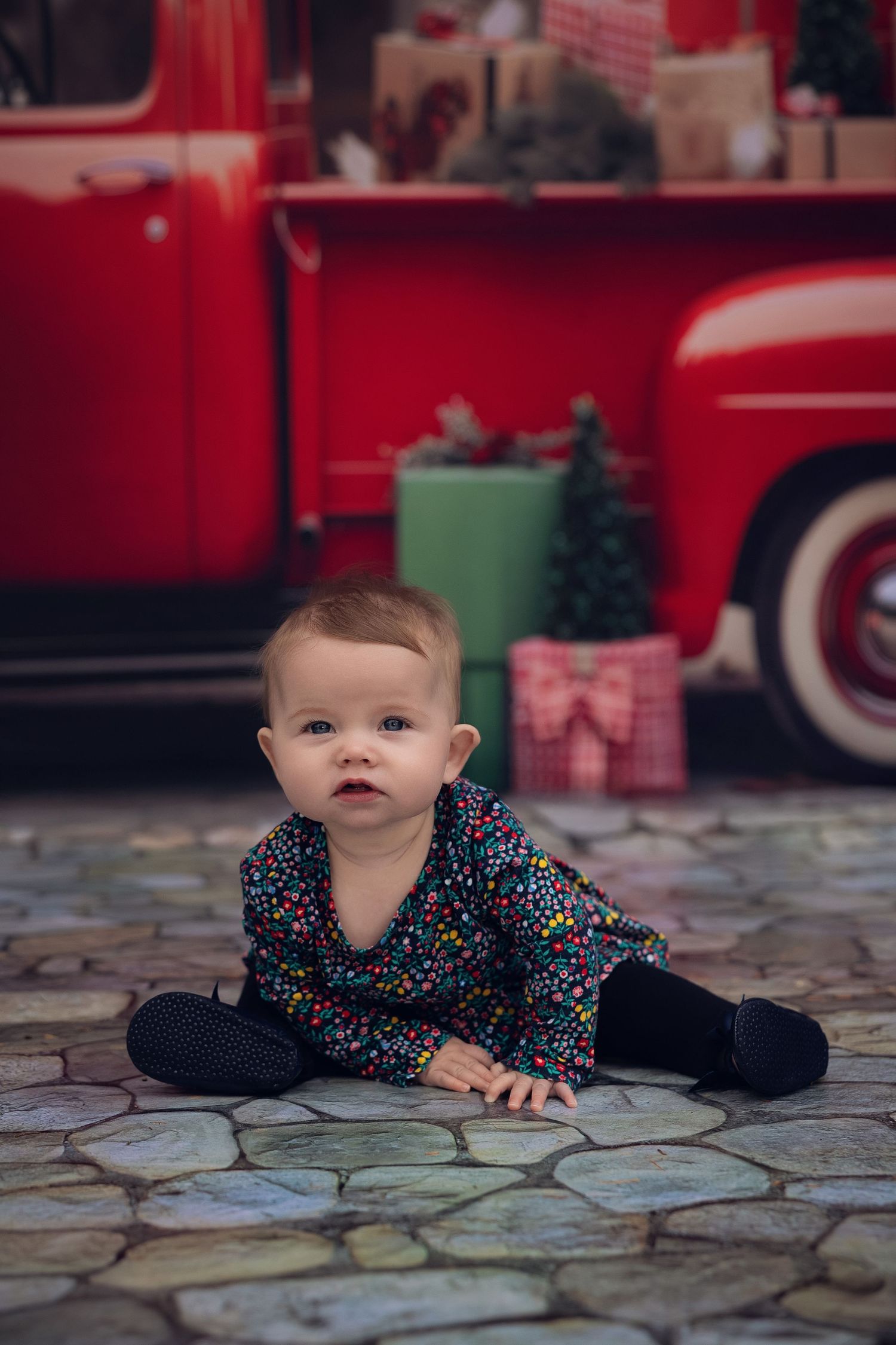 Baby in floral outfit sits on stone floor before a vintage red Christmas truck with gifts and a small tree.