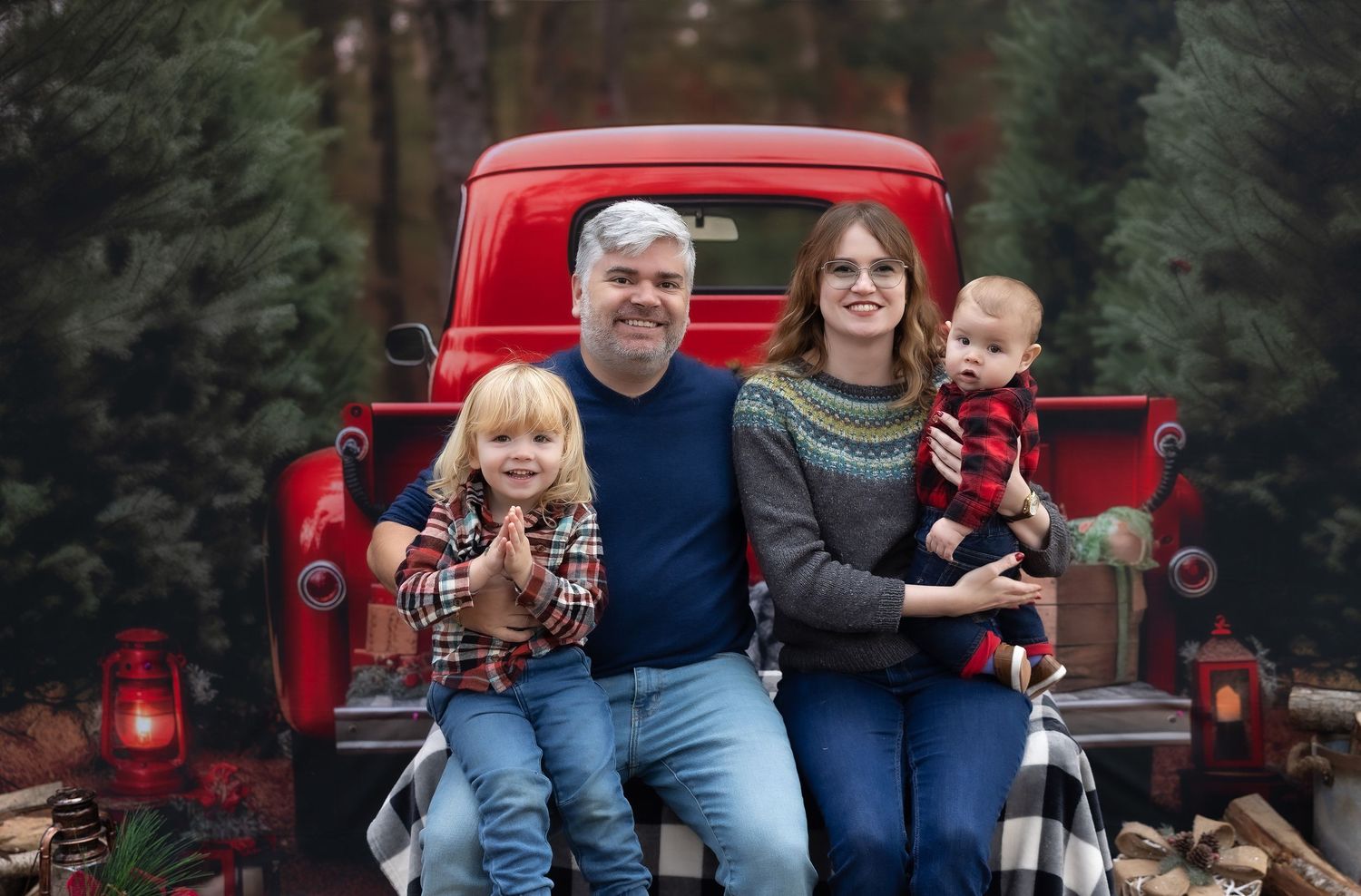 Family of four posing on red vintage truck tailgate surrounded by pine trees and festive holiday decor.