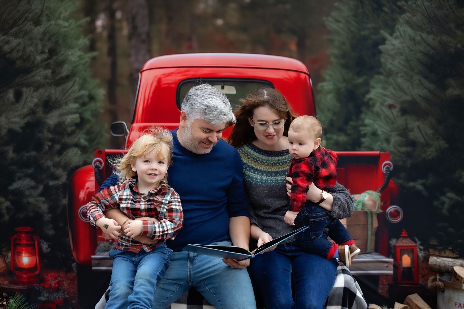 Family of four sitting on red truck bed with Christmas trees, reading a book together in holiday outfits.